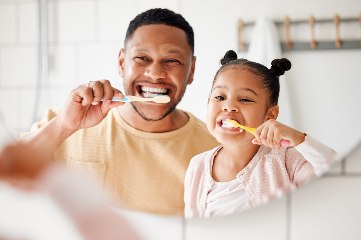 a father and young daughter brush their teeth together in the mirror