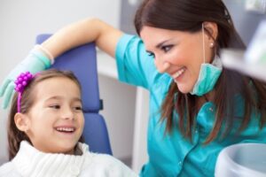 A woman dentist with blue scrubs smiling at a girl in her exam chair.