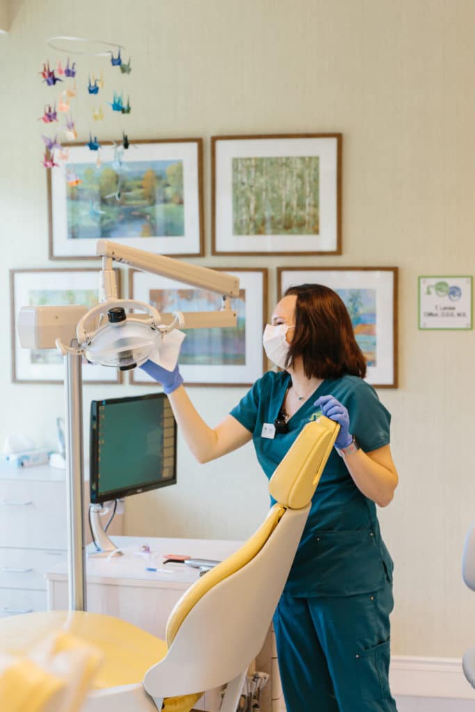 A dental assistant in teal scrubs and a mask adjusts an exam light.