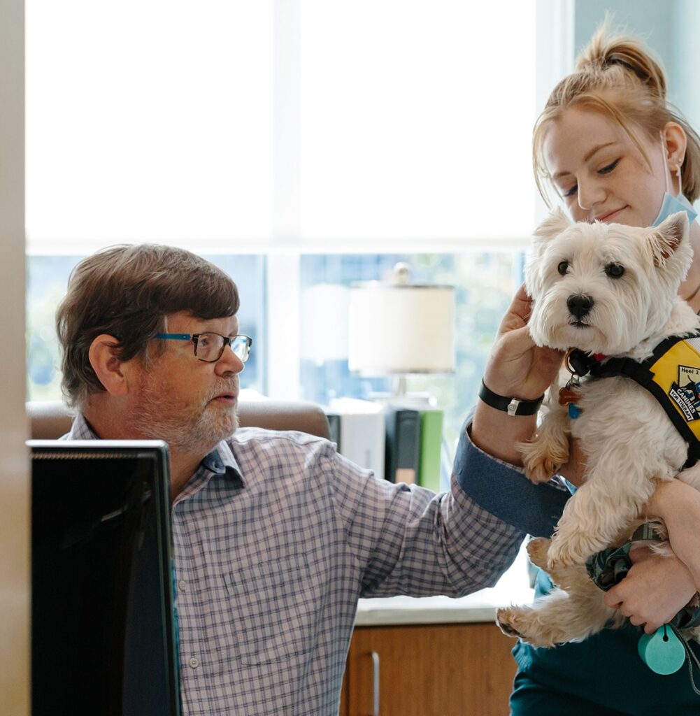 Pediatric dentist Dr. Chuck at his desk petting Sugar, the office therapy dog! Sugar is being held by his handler, a young adult in blue scrubs.