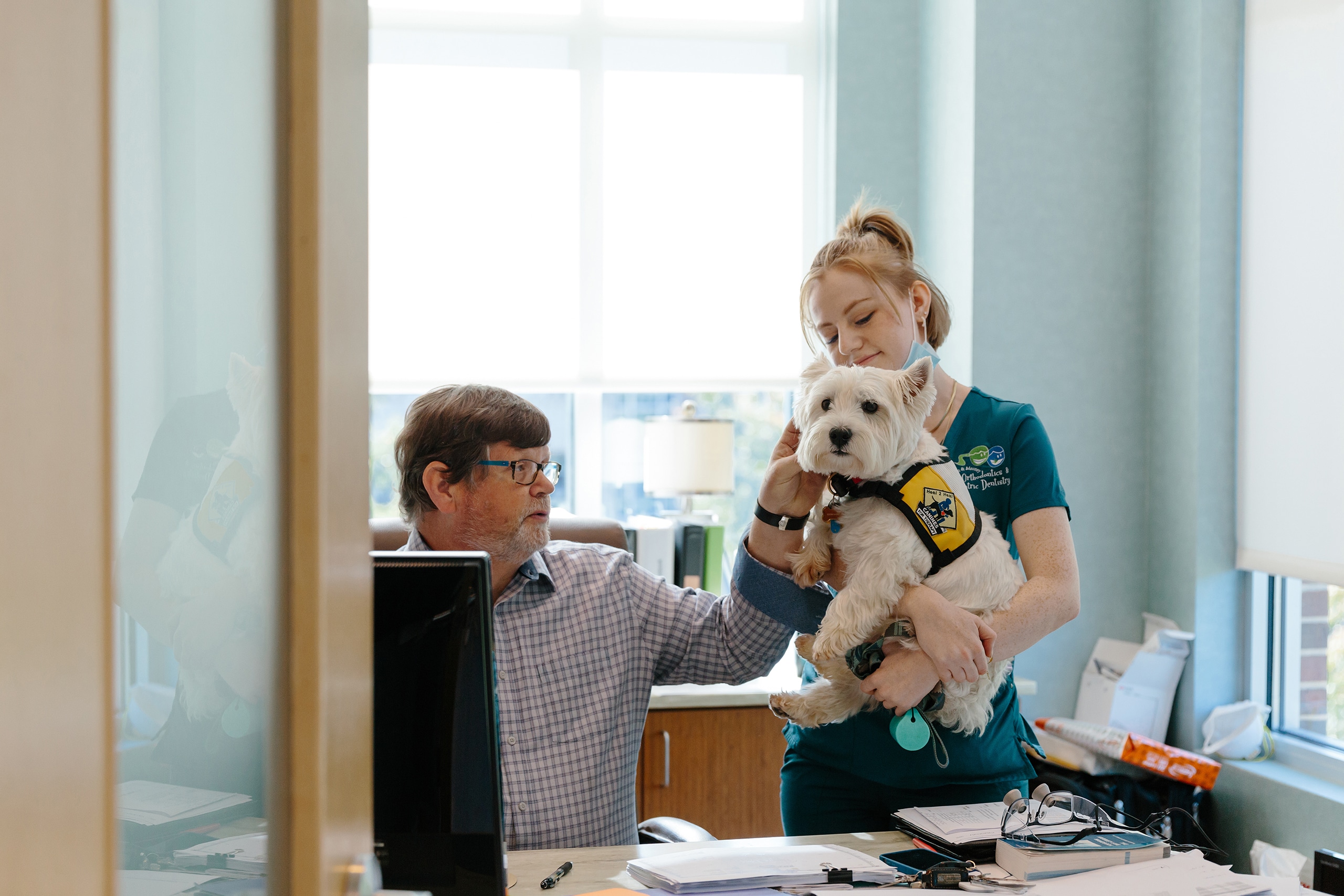 A man at his desk petting a dog that is held by a woman in blue scrubs.