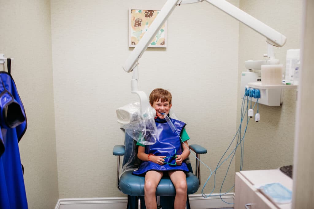 A young child sits in an xray room, in the midst of getting a classic x-ray with bitewings at the pediatric dentist. He's also, impressively, smiling at the photographer behind this photo.