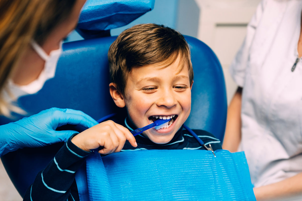 A young boy smiles while brushing his teeth during a dental visit, guided by a dental professional.