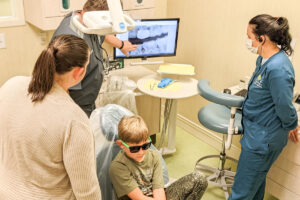 A dentist shows dental X-rays on a monitor to a parent and child wearing sunglasses during a checkup.