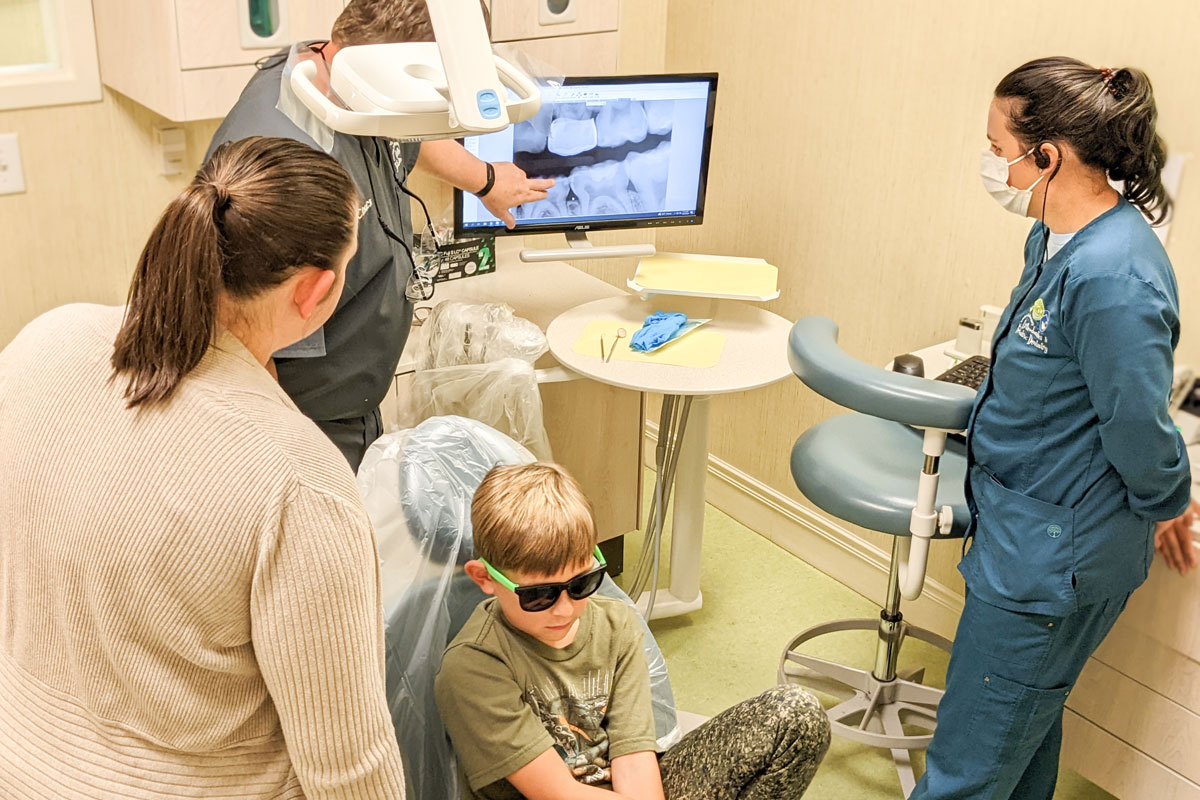 A dentist shows dental X-rays on a monitor to a parent and child wearing sunglasses during a checkup.