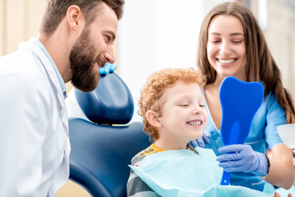 A smiling young child sits in a dental chair holding a blue mirror while a dentist and a dental assistant stand beside him, happily watching his reaction.