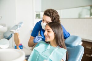 A dental professional wearing gloves and a face mask points at a young girl’s teeth while the girl, sitting in a dental chair with braces and a bib, smiles and looks into a small handheld mirror.