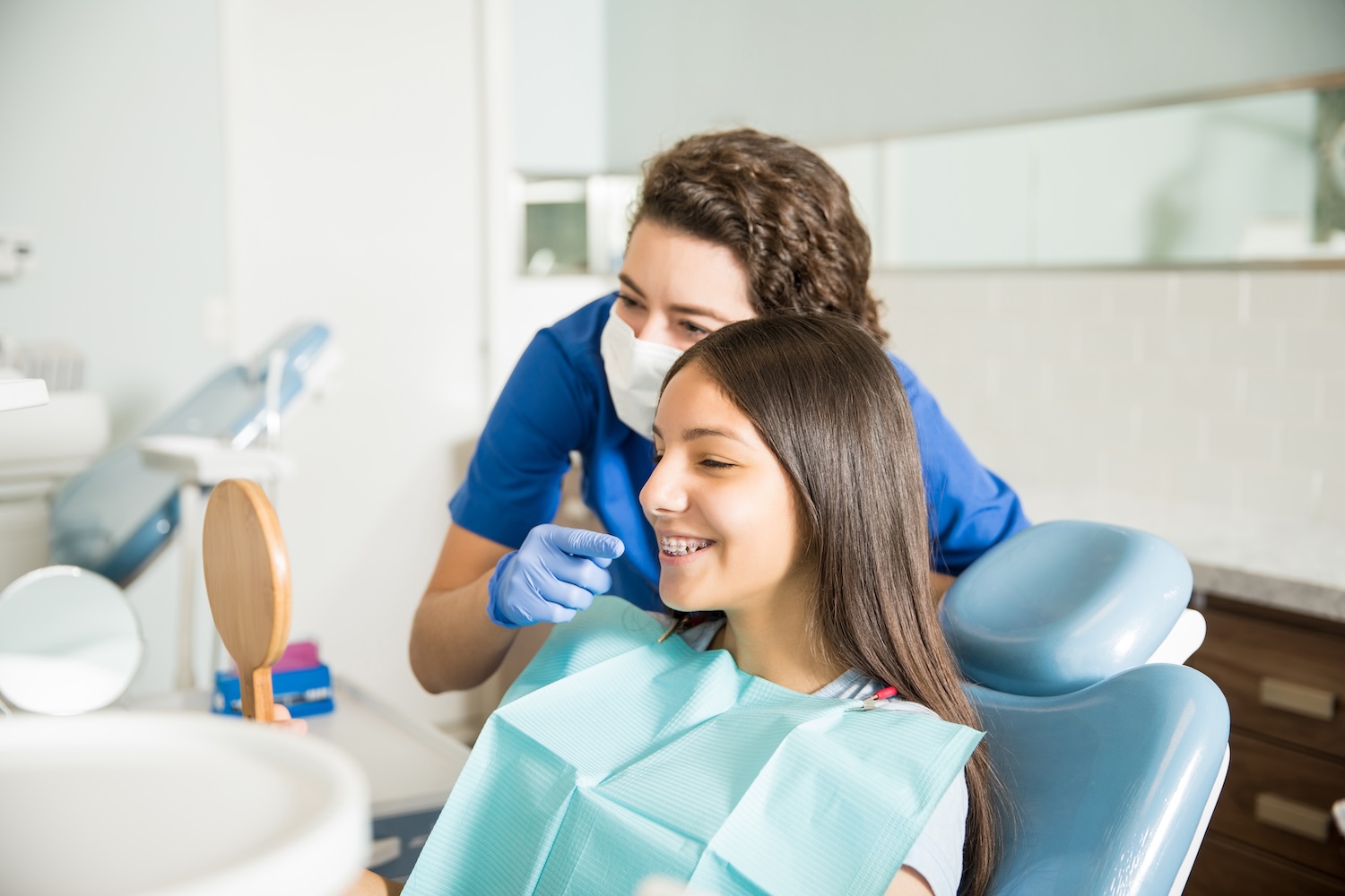 A dental professional wearing gloves and a face mask points at a young girl’s teeth while the girl, sitting in a dental chair with braces and a bib, smiles and looks into a small handheld mirror.